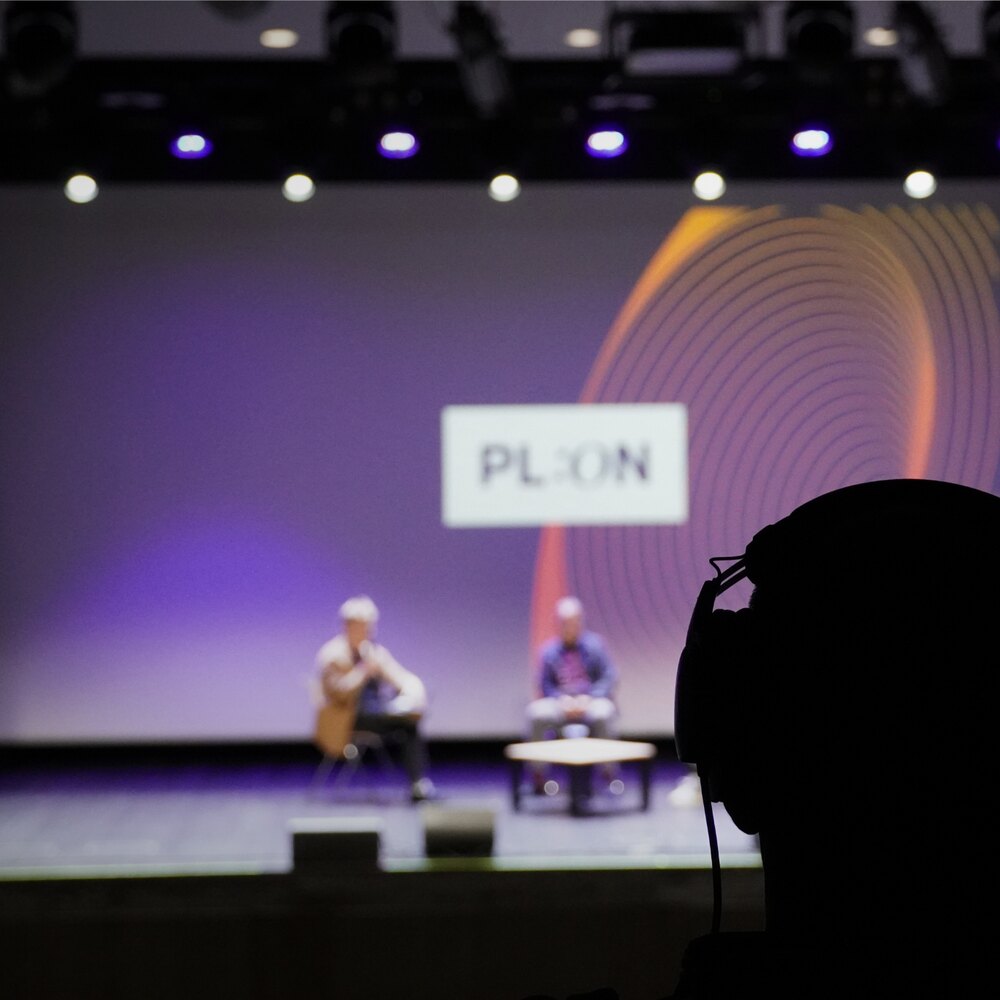 Scena festiwalu PL:ON w Olsztynie w 2025 roku — dwoje rozmówców podczas panelu, za nimi plansza z logo PL:ON, w kontrze sylwetka kamery.